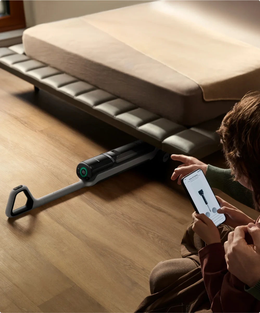 Dreame H15 Pro Heat incliné sous un canapé, utilisateur à côté avec un téléphone